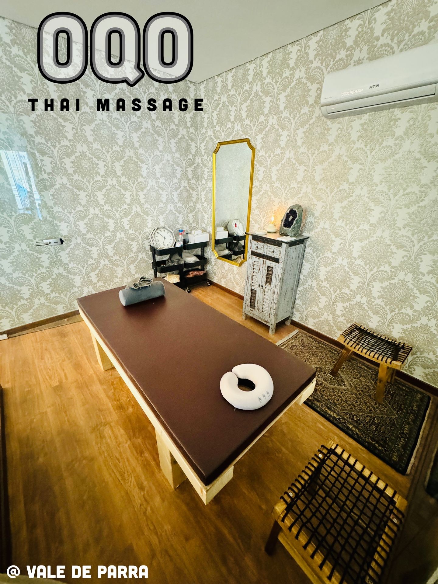 Thai massage room with a massage table, decorative mirror, and chairs, featuring floral wallpaper.